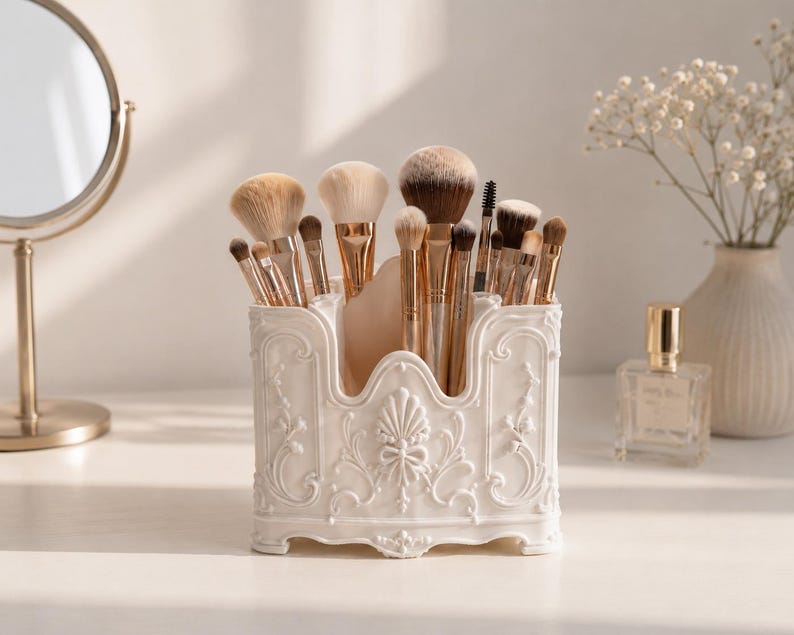 May include: A white plastic makeup brush holder featuring an ornate, raised relief design with floral and scroll motifs. The rectangular container has a scalloped top edge and holds a collection of various makeup brushes with gold-toned ferrules.