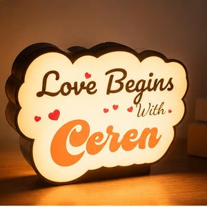 May include: A cloud-shaped LED lamp with a warm white glow, featuring the dark brown text "Love Begins With" and the orange text "Ceren" in a bold font, accented by several small red hearts printed on the front surface of the light.
