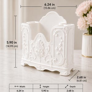 May include: A white desk organizer featuring ornate relief detailing, including a central shell and bow motif. It measures 6.24 inches in width, 5.90 inches in height, and 2.68 inches in depth, with multiple compartments for storage and a raised back panel.