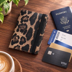 May include: A rectangular passport holder featuring a black and metallic gold leopard print pattern with a vertical black elastic closure strap. Beside it lies a blue United States passport, a boarding pass for Tel Aviv to New York, and a black Visa card.