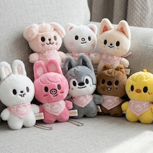 May include: A collection of plush animal keychains in various colors, including white, pink, gray, and yellow. Each keychain features a unique animal design, such as a bunny, pig, cat, and chick, and wears a pink scarf. They are arranged on a gray sofa.