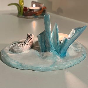 May include: A handmade ceramic candle holder featuring a seal figurine on a light blue base. The holder includes blue ice-like structures and a small candle. Another candle holder is visible in the background.