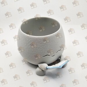 May include: A grey ceramic planter in the shape of a character, holding a small blue book. The planter has a textured surface and a wide opening. The character has closed eyes and a smiling mouth.