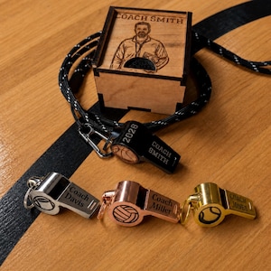 May include: A collection of four metal whistles in black, silver, rose gold, and gold finishes, each engraved with a name and sports ball icon. A black lanyard and a small wooden box featuring an engraved portrait and the text COACH SMITH.