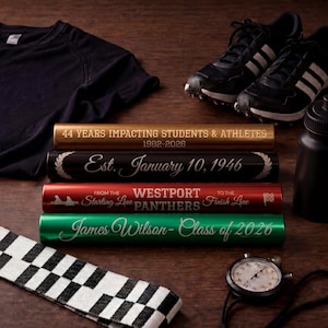 May include: Four cylindrical relay batons in gold, black, red, and green with custom engravings. The gold baton reads 44 YEARS IMPACTING STUDENTS & ATHLETES 1982-2026. The black baton reads EST. January 10, 1946. The red baton reads FROM THE STARTING LINE WESTPORT PANTHERS TO THE FINISH LINE. The green baton reads James Wilson- Class of 2026.