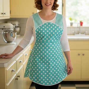 May include: A turquoise polka dot apron with white trim and straps. The apron features a classic design with a full skirt and a bib top. The white polka dots are evenly spaced across the turquoise fabric. The apron is worn over a white long-sleeved shirt.