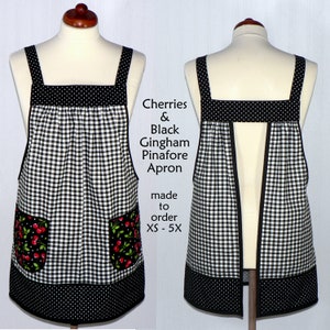 May include: Black and white gingham pinafore apron with cherry print pockets. The apron has a black and white polka dot trim and a split back. "Cherries & Black Gingham Pinafore Apron made to order XS - 5X" is written on the apron.