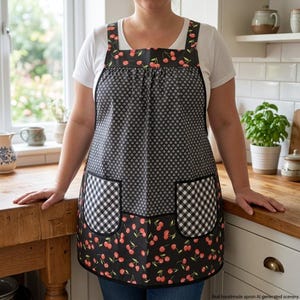 May include: A black apron featuring a cherry print on the top and bottom sections, with a central panel of small white geometric shapes on a black background. The apron includes two front pockets with a black and white gingham check pattern.