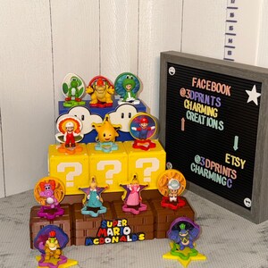 May include: A collection of Super Mario McDonald's toys, featuring various characters like Mario, Princess Peach, and Yoshi. The toys are displayed on yellow question mark blocks and brown platforms. A black letter board with social media handles is in the background.
