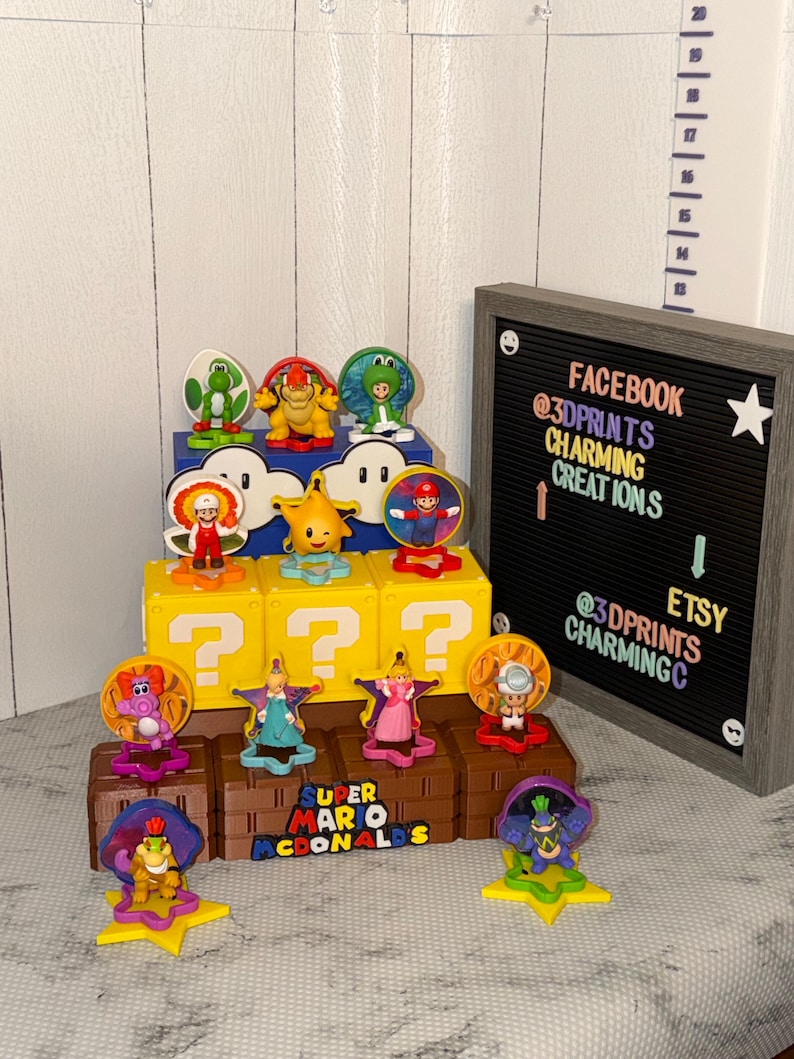 May include: A collection of Super Mario McDonald's toys, featuring various characters like Mario, Princess Peach, and Yoshi. The toys are displayed on yellow question mark blocks and brown platforms. A black letter board with social media handles is in the background.