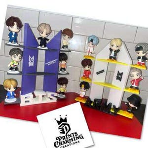 May include: A display of miniature figurines of a musical group, arranged on white and purple shelves. The figurines are of various colors and styles. The display sits on a red surface, with a white card featuring a logo in the foreground.