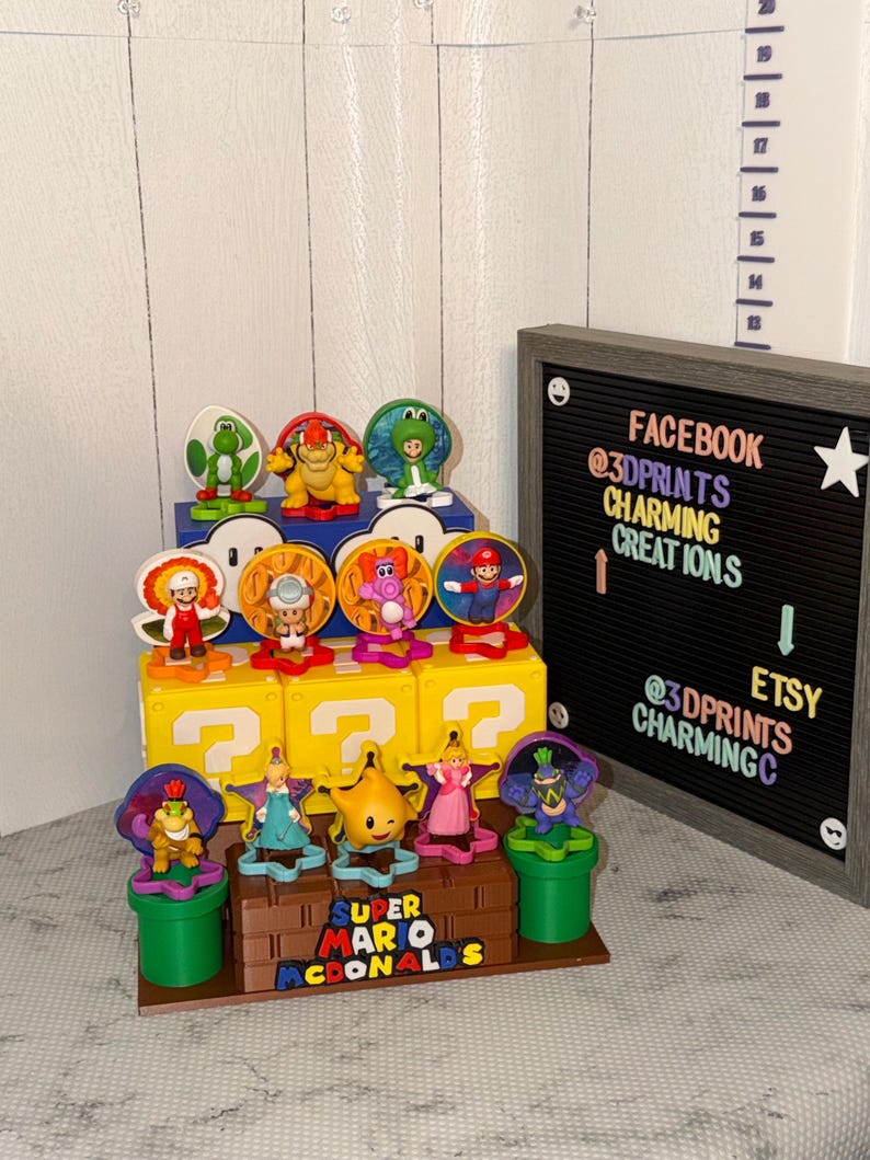 May include: A Super Mario display featuring various character figurines. The display includes yellow question mark blocks, green pipes, and a sign that reads "Super Mario McDonalds". The sign also includes Etsy and Facebook information.