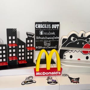May include: A black city skyline display shelf with red accents, a black sign with white text reading CHECKUS OUT Facebook 3DPrintsCharmingCreation Etsy @3DPrintsCharmingC, a yellow McDonald's logo, a white Hello Kitty shelf, and three character keychains.