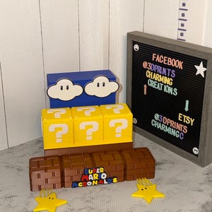 May include: A display of 3D-printed Super Mario-themed items. Includes a blue box with cloud designs, yellow question mark blocks, brown brick blocks, and yellow star-shaped objects. A letter board displays social media handles.