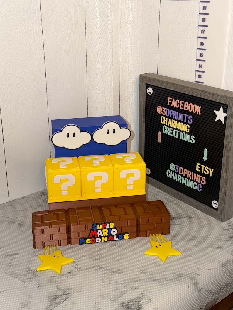 May include: A display of 3D-printed Super Mario-themed items. Includes a blue box with cloud designs, yellow question mark blocks, brown brick blocks, and yellow star-shaped objects. A letter board displays social media handles.