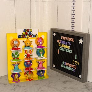 K&ouml;nnte beinhalten: Ein gelbes Display mit dem Text "Super Mario McDonalds" zeigt verschiedene bunte Figuren. Rechts befindet sich eine gerahmte Buchstaben-Tafel mit Social-Media-Handles und einem Stern. Im Hintergrund ist ein Lineal zu sehen.