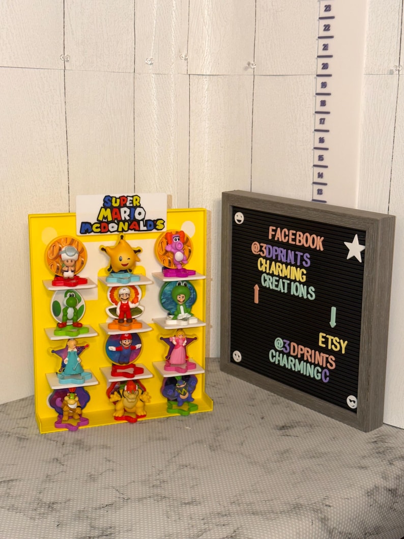 K&ouml;nnte beinhalten: Ein gelbes Display mit dem Text "Super Mario McDonalds" zeigt verschiedene bunte Figuren. Rechts befindet sich eine gerahmte Buchstaben-Tafel mit Social-Media-Handles und einem Stern. Im Hintergrund ist ein Lineal zu sehen.