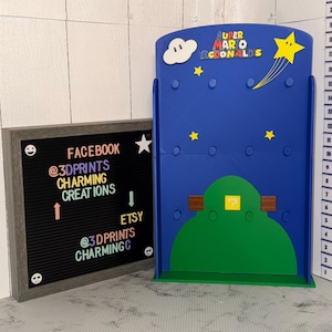 May include: A blue Super Mario-themed display stand with a green hill, yellow stars, and a cloud. The stand has the text "SUPER MARIO MCDONALDS". A framed letter board displays social media information: "FACEBOOK @3DPRINTS CHARMING CREATIONS" and "ETSY @3DPRINTS CHARMINGC".