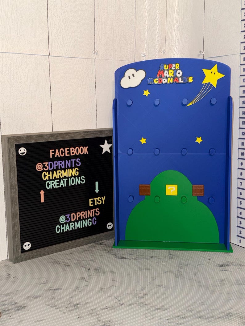 May include: A blue Super Mario-themed display stand with a green hill, yellow stars, and a cloud. The stand has the text "SUPER MARIO MCDONALDS". A framed letter board displays social media information: "FACEBOOK @3DPRINTS CHARMING CREATIONS" and "ETSY @3DPRINTS CHARMINGC".