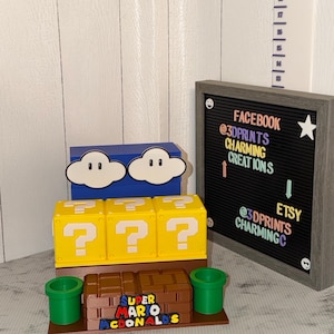 May include: A display of 3D-printed Super Mario-themed items. Includes yellow question mark boxes, a blue cloud box, a brown box with the text "SUPER MARIO MCDONALDS", and green pipes. A framed sign displays social media information.