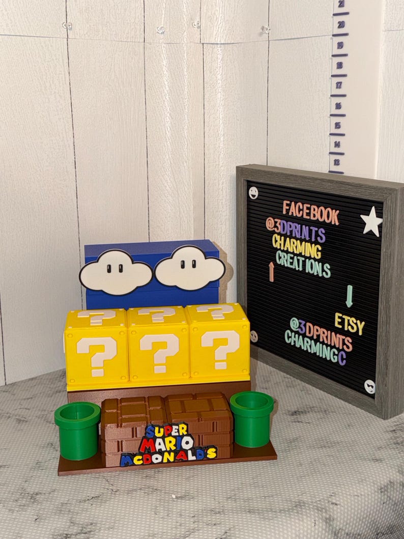 May include: A display of 3D-printed Super Mario-themed items. Includes yellow question mark boxes, a blue cloud box, a brown box with the text "SUPER MARIO MCDONALDS", and green pipes. A framed sign displays social media information.
