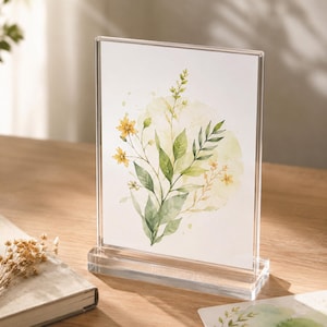 Könnte beinhalten: Transparenter Acryl-Bilderrahmen mit einem Aquarell-Blumendruck. Das Kunstwerk zeigt gelbe Blumen und grüne Blätter vor einem weichen gelben Hintergrund. Der Rahmen ist rechteckig mit einem transparenten Sockel.