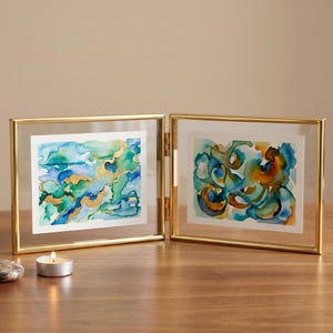 May include: A gold-framed, two-panel display featuring watercolor art. The left panel showcases a blue, green, and gold abstract design. The right panel displays a similar abstract piece in blue, green, and orange. A small candle and a stone are on the wooden surface.