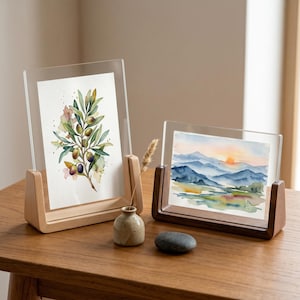 Könnte beinhalten: Zwei klare Acrylrahmen mit Holzsockeln, die Aquarelldrucke enthalten. Der vertikale Rahmen zeigt einen Olivenzweig mit grünen und violetten Oliven. Der horizontale Rahmen zeigt eine Landschaft mit blauen Bergen bei Sonnenuntergang und einen Stein.