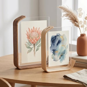 May include: Two clear acrylic and wood picture frames. One frame has a watercolor painting of a pink flower, the other has a blue and gold abstract painting. The frames are on a wooden table.