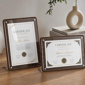 Pode incluir: Dois certificados emoldurados com bordas escuras de grão de madeira e frentes de acrílico transparente. Os certificados dizem "Certificado de Conquista" e "Certificado de Apreciação". Cada moldura tem um suporte de metal.