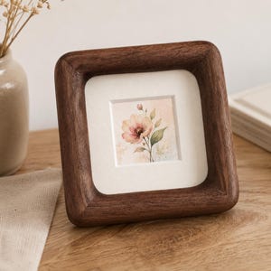 May include: A square, wooden picture frame with a watercolour floral print. The frame is made of dark brown wood and has rounded corners. The artwork features a pink and peach flower with green leaves.