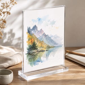 Può includere: Una cornice fotografica in acrilico trasparente che mostra un dipinto di paesaggio ad acquerello di montagne, un lago e alberi. L'opera d'arte presenta sfumature di blu, verde, giallo e arancione. La cornice ha una base rettangolare e un orientamento verticale.