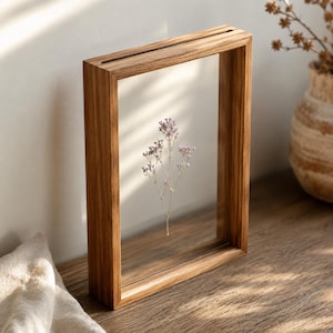 May include: A wooden rectangular frame with a clear glass centre, displaying a delicate sprig of dried, light purple flowers. The frame has a natural wood grain finish and a slot at the top. The arrangement is displayed on a wooden surface.