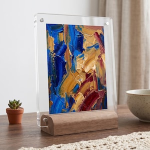Könnte beinhalten: Abstraktes Gemälde in einem Rahmen mit Holzsockel. Das Kunstwerk zeigt dicke Pinselstriche in Blau, Gold und Rot. Der Rahmen ist aus klarem Acryl, der Sockel aus hellem Holz. Eine kleine Sukkulente in einem Terrakotta-Topf ist zu sehen.