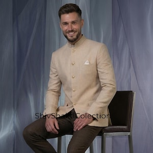 Puede incluir: Un hombre con una chaqueta beige de cuello mao, pantalones marrones y zapatos negros, sentado en una silla marrón con estructura plateada. El fondo es una cortina azul claro. La chaqueta tiene un pañuelo.