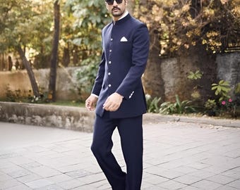 May include: A navy blue suit with a mandarin collar jacket and matching trousers. The jacket has a row of buttons and a pocket square. The person is wearing sunglasses and brown leather shoes. The background features trees and a paved walkway.