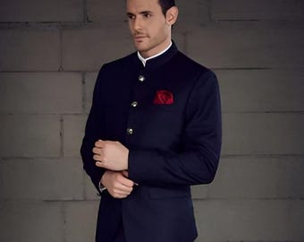May include: A navy blue suit with a mandarin collar, gold buttons, and a red pocket square. The suit includes a matching jacket and trousers. The person is wearing black dress shoes. The background is a gray brick wall.