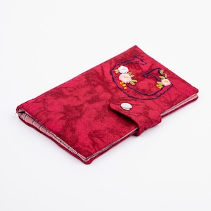 Może przedstawiać: Czerwony, materiałowy portfel z haftowanym kwiatowym wzorem. Portfel posiada zapięcie na guzik i wzorzyste wnętrze. Haftowany wzór przedstawia literę "G" z kwiatami w odcieniach bieli, różu i żółci.