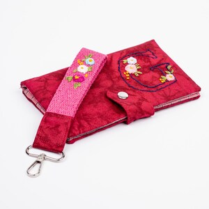Może przedstawiać: Czerwony materiałowy portfel z pasującym różowym paskiem i srebrnym zapięciem. Portfel i pasek zdobione są haftowanymi kwiatami. Litera "G" jest wyhaftowana na przodzie portfela.