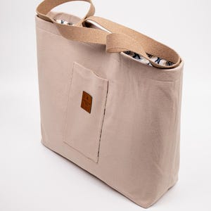 May include: A beige tote bag with a rectangular shape and a front pocket. The bag has tan-colored straps and a patterned interior. A small, brown square label is sewn onto the front pocket. The bag is made of a sturdy material.