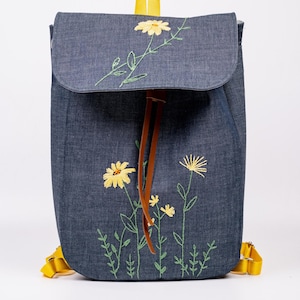 Op de afbeelding: Denim rugzak met gele madeliefjes borduursel. De rugzak heeft een klepsluiting, gele bandjes en een trekkoordsluiting. Het bloemmotief omvat gele bloemen en groene stelen.