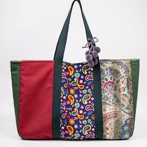 May include: A large tote bag with a patchwork design. The bag features panels of red, green, and paisley fabric in various colours. Dark green handles and a pom-pom charm complete the design. The bag is designed for carrying items.