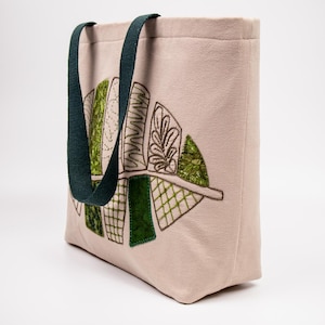 May include: Beige tote bag with dark green handles and an embroidered leaf design. The leaf design features various shades of green and white stitching. The bag is made of a textured fabric, ideal for carrying everyday items.