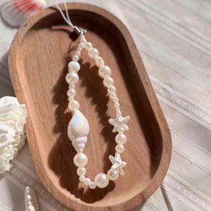 May include: A beaded phone charm featuring a pearlescent seashell and white starfish accents. The charm is strung with white faux pearls of varying sizes. The charm is displayed on a wooden tray, suggesting a beach or ocean theme.