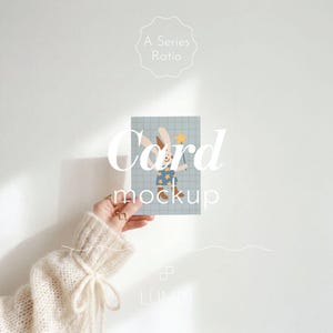 Puede incluir: Una maqueta de tarjeta con un conejito de dibujos animados sosteniendo una varita de estrella, sobre un fondo de cuadrícula azul claro. La tarjeta se sostiene frente a una pared blanca. Las palabras "Card mockup" se muestran en la tarjeta.