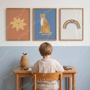 K&ouml;nnte beinhalten: Drei gerahmte Kunstdrucke mit Cartoon-Illustrationen. Die Drucke zeigen eine l&auml;chelnde Sonne, einen Leoparden und einen Regenbogen. Die Rahmen sind hellbraun. Der Hintergrund ist eine hellblaue und wei&szlig;e Wand.