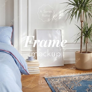 May include: A white frame mockup with a grid pattern, leaning against a white wall. A blue bedspread with red trim is visible on the left. A small blue and white striped cup sits on a stack of books. A potted plant and a blue rug complete the scene.