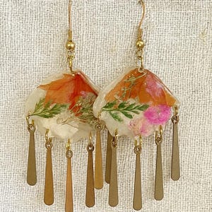 Puede incluir: Pendientes hechos a mano con un diseño en forma de abanico, que incorporan flores secas en naranja, rosa y verde. Los pendientes tienen detalles de metal dorado y colgantes en forma de lágrima. Miden aproximadamente 5 cm de largo.