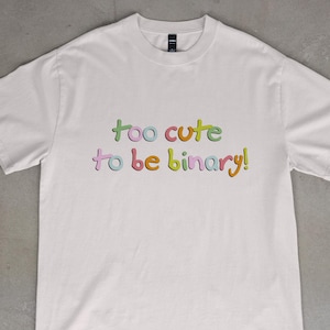 May include: White t-shirt with the text "too cute to be binary!" in colorful, bubble-style lettering. The shirt is made of a soft material and has short sleeves. The text is in shades of green, pink, blue, yellow, and orange.