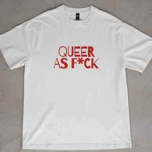 May include: White t-shirt with the red text "QUEER AS F*CK" printed across the front. The shirt is a crew neck style and appears to be made of a soft material. The shirt is displayed on a gray surface.
